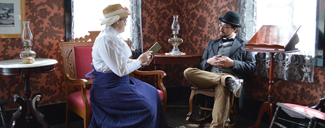 A woman and man sit together dressed in old-fashioned clothing.