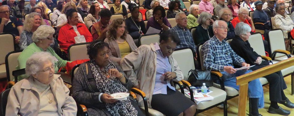 A large group of senior citizens sit and look toward a stage.