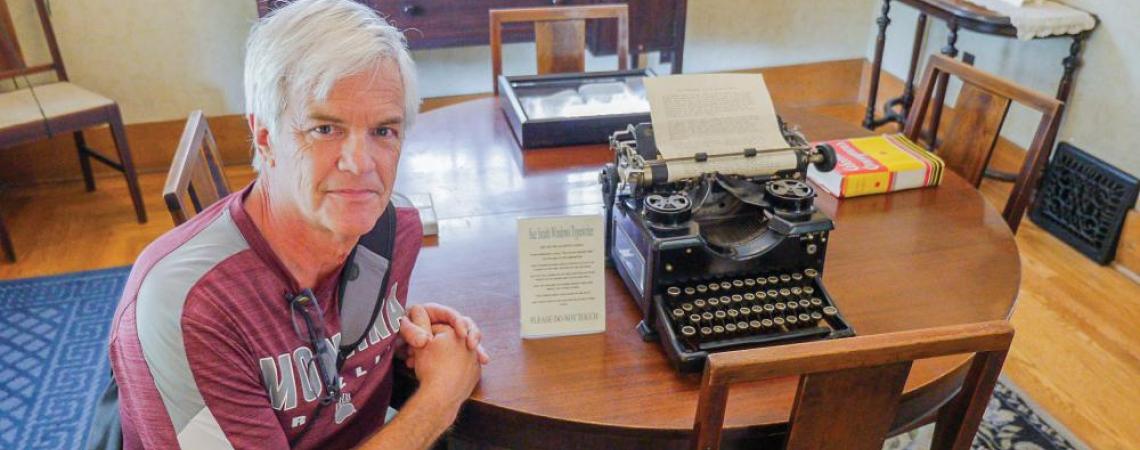 Eric Dentler poses at a table with the typewriter used to write “The Big Book.” In the background, an original copy is encased in glass. 