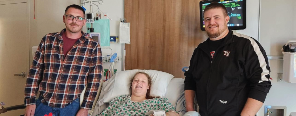 A young girl in a hospital bed beside the two men who saved her life