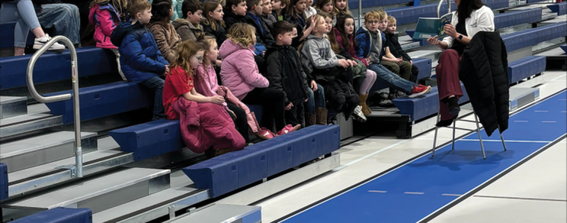 A woman reading a book to a group of young children in a school gymnasium